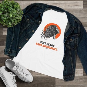 May include: A white t-shirt featuring a graphic of a profile silhouette with roses in the hair against an orange circle. Below the graphic, the text reads: SOFT HEART. HARD BOUNDARIES. The shirt is layered under a dark blue denim jacket.
