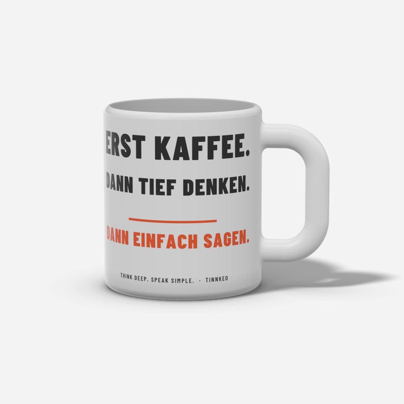 Op de afbeelding: Een witte keramische mok met een groot handvat, voorzien van zwarte tekst die luidt ERST KAFFEE. DANN TIEF DENKEN. en oranje tekst die luidt DANN EINFACH SAGEN. Daaronder staat in kleinere letters THINK DEEP. SPEAK SIMPLE. &middot; TINNKEO.