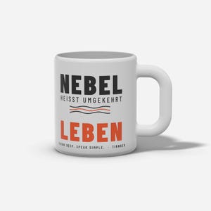 Puede incluir: Una taza de cer&aacute;mica blanca con el texto negro NEBEL HEISST UMGEKEHRT, seguido de dos l&iacute;neas onduladas en negro y naranja. Debajo, la palabra LEBEN est&aacute; impresa en letras grandes de color naranja, con el texto THINK DEEP. SPEAK SIMPLE. TINNKEO.