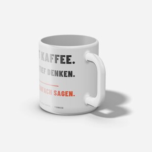 Op de afbeelding: Een witte keramische mok met zwarte tekst die MIT KAFFEE. TIEF DENKEN. leest en rode tekst die EINFACH SAGEN. leest. Een horizontale rode lijn scheidt de zinnen en de kleine tekst onderaan leest KEEP IT SIMPLE. TINNKEO.