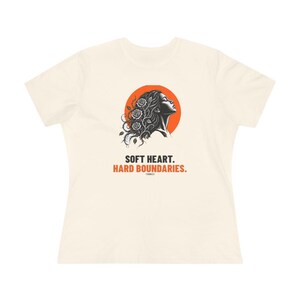 May include: A cream-colored short-sleeve t-shirt featuring a graphic of a profile silhouette with roses in the hair against an orange circle. Below the graphic, the text reads: SOFT HEART. HARD BOUNDARIES. The design is centered on the chest area.