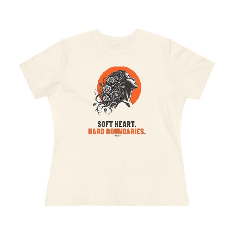 May include: A cream-colored short-sleeve t-shirt featuring a graphic of a profile silhouette with roses in the hair against an orange circle. Below the graphic, the text reads: SOFT HEART. HARD BOUNDARIES. The design is centered on the chest area.