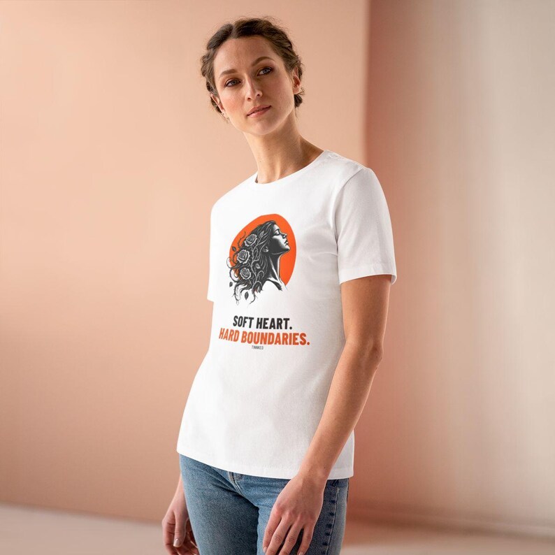 May include: A white short-sleeved t-shirt featuring a graphic of a person's profile with floral hair against an orange circle. Below the graphic, the text reads "SOFT HEART. HARD BOUNDARIES." in black and orange capital letters on the chest.