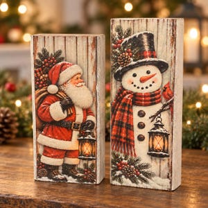 Puede incluir: Dos bloques de madera rectangulares con ilustraciones navideñas. Un bloque muestra a Santa Claus con un saco y una linterna, mientras que el otro muestra un muñeco de nieve con sombrero de copa y bufanda a cuadros, sosteniendo una linterna.