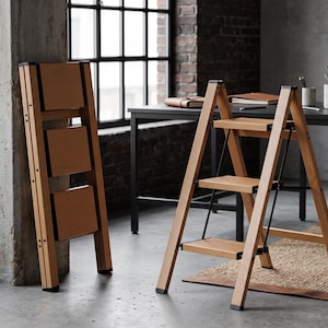 May include: A three-step folding ladder made of light-toned wood with black metal hinges and supports. One ladder is shown in a fully open, standing position, while a second identical ladder is shown folded flat and leaning against a pillar.