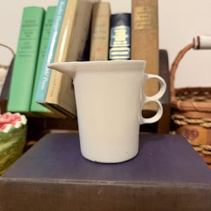 May include: A white ceramic creamer with a speckled finish, featuring a pointed pouring spout and a unique handle design consisting of two stacked circular loops. The vessel has a cylindrical body and a smooth, matte-textured surface.
