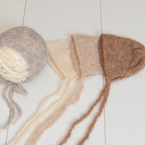 May include: Three knitted baby bonnets in shades of beige, brown, and gray. The bonnets have ties that can be tied under the chin.