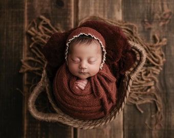 May include: A newborn baby wrapped in a rust-colored knit blanket and bonnet with a white lace trim, nestled in a woven basket. The basket sits on a rustic wooden surface, creating a warm and cozy scene.