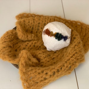 May include: A mustard yellow knitted blanket with a white foam ball topped with a colourful flower crown. The crown is made of fabric flowers in shades of orange, yellow, green, and blue.