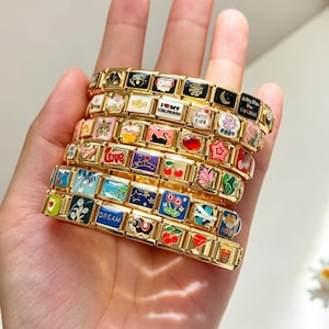 Könnte beinhalten: Ein Stapel goldfarbener Gliederarmbänder mit verschiedenen bunten Emaille-Anhängern. Die Anhänger zeigen diverse Motive wie ein Spinnennetz, einen Mond, Kirschen, einen Schmetterling, einen Bubble Tea sowie Texte wie I MY GIRLFRIEND, LOVE und DREAM.