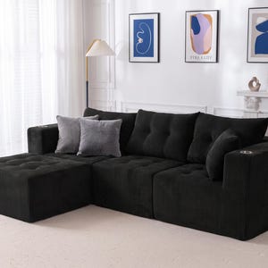 May include: A black corduroy sectional sofa featuring a chaise lounge, tufted seat cushions, and integrated cup holders in the armrests. The sofa is accented with two grey square throw pillows and includes a matching black bolster pillow.