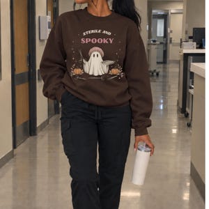May include: A dark brown crewneck sweatshirt featuring a graphic of a ghost wearing a surgical cap, holding surgical instruments and a tray. The text reads STERILE AND SPOOKY, surrounded by small pumpkins, stars, and medical tools.