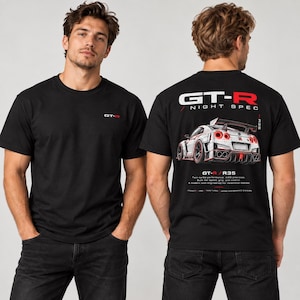 以下が含まれることがあります： 胸元に小さな赤と白のGT-Rロゴが入った黒いTシャツ。背面には、赤いテールライトを備えた白いスポーツカーの大きなグラフィック、GT-R NIGHT SPECというテキスト、および白い文字で詳細な技術仕様がプリントされています。