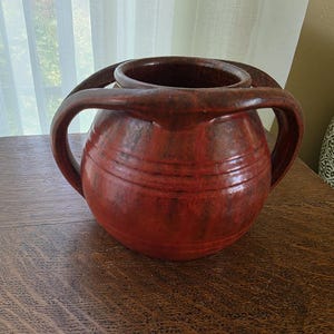 May include: A round, dark red ceramic vessel with a wide opening and two large, curved handles extending from the rim to the midsection. The surface features a mottled, textured finish with subtle horizontal ridges encircling the body.