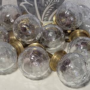 Puede incluir: Un conjunto de pomos esféricos para armarios con superficies de cristal craquelado transparente y bases de metal en tono latón. Cada pomo tiene una parte superior de cristal texturizado con un patrón de red y un tornillo roscado.
