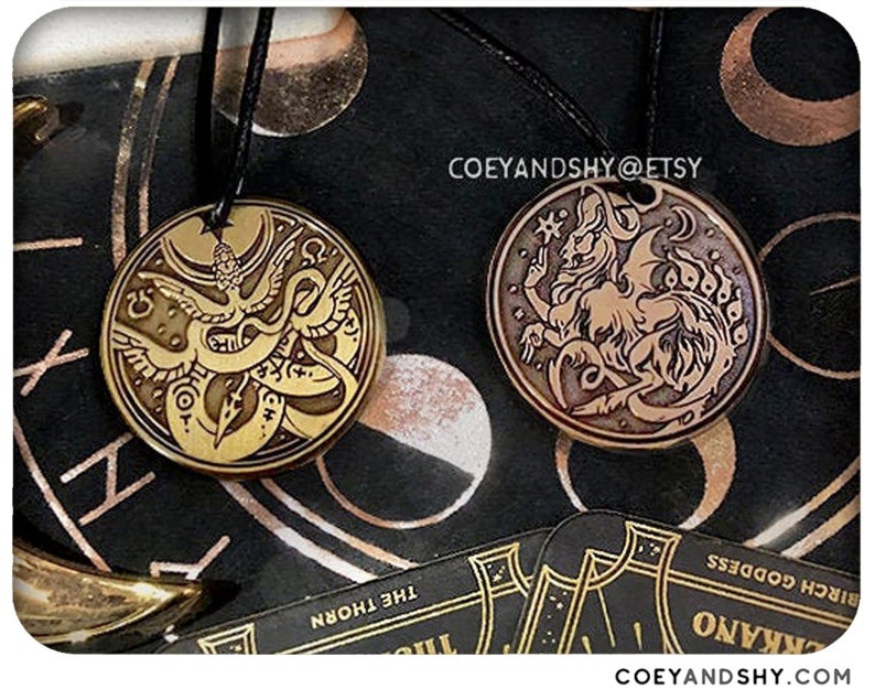 Puede incluir: Dos colgantes de metal dorado en cordones negros. El colgante de la izquierda presenta un p&aacute;jaro estilizado con una serpiente envuelta a su alrededor. El colgante de la derecha presenta una criatura estilizada con alas y cola. Ambos colgantes tienen un fondo marr&oacute;n oscuro.