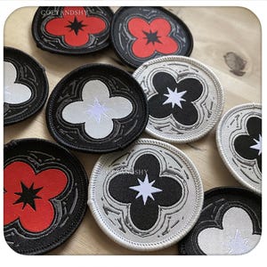 Puede incluir: Una colección de parches bordados circulares con un diseño de flor de cuatro pétalos y una estrella central. Los parches vienen en negro con flores rojas, negro con flores blancas y blanco con flores negras, todos con el texto COZYANDSHY.