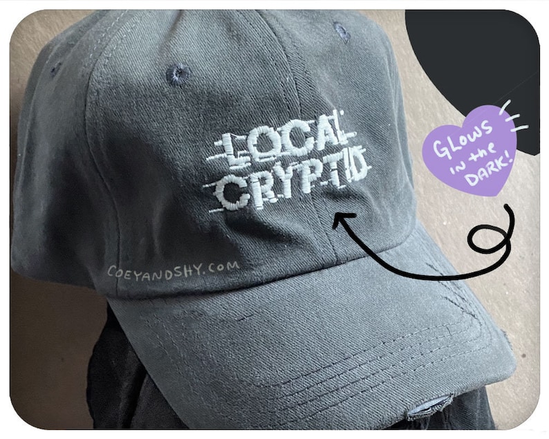 May include: A gray baseball cap with the text "LOCAL CRYPTID" embroidered in white on the front. The text is in a pixelated font. The cap has a white embroidered logo on the side that says "COEYANDSHY.COM". A purple heart with the text "Glows in the DARK!" is in the upper right corner of the image.