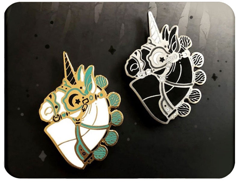 May include: Two enamel pins featuring a unicorn head with a crescent moon and star on its forehead. One pin is white with teal accents and the other is black with silver accents.