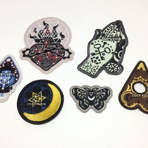 Puede incluir: Seis parches bordados para planchar con un esquema de colores negro, blanco y dorado. Los parches presentan varios dise&ntilde;os, incluyendo un coraz&oacute;n, una mano, una luna creciente, una mariposa y un tri&aacute;ngulo. El texto "COEY KUHN" aparece en cada parche.