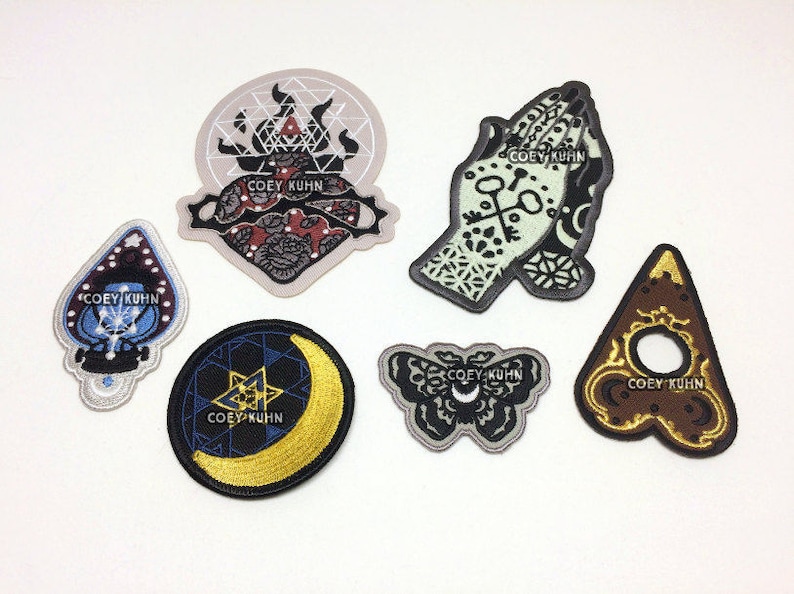 Puede incluir: Seis parches bordados para planchar con un esquema de colores negro, blanco y dorado. Los parches presentan varios dise&ntilde;os, incluyendo un coraz&oacute;n, una mano, una luna creciente, una mariposa y un tri&aacute;ngulo. El texto "COEY KUHN" aparece en cada parche.