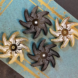 May include: Four enamel pins in the shape of a starburst with a central star. Two pins are black with white stars and two pins are gold with black stars.