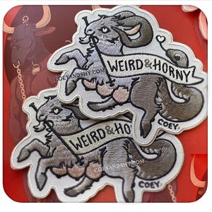 May include: Embroidered patches featuring a gray ram with large horns, holding a banner that reads "WEIRD & HORNY." The patches have a white border and are stacked on a red background. The website "COEYANDSHY.COM" is also visible.