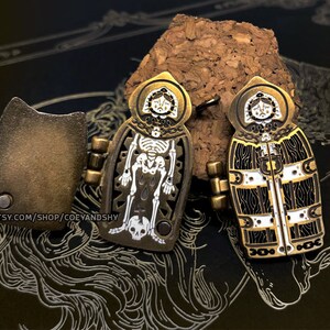 May include: Two gold-toned metal pins with a skeleton and a woman's face in the center. The pins are attached to a cork surface. The skeleton pin has a cross and a skull at the bottom. The woman's face pin has a white and black patterned design.
