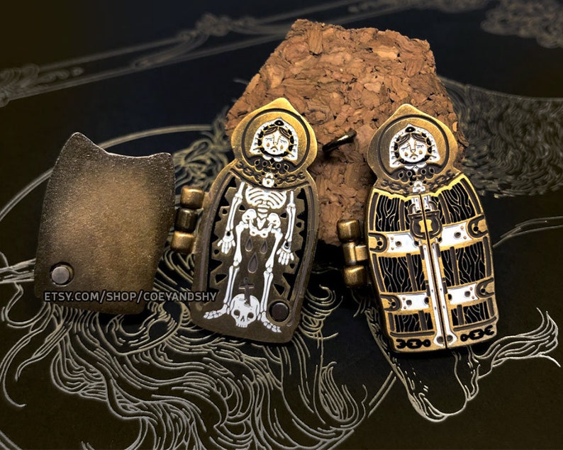 May include: Two gold-toned metal pins with a skeleton and a woman's face in the center. The pins are attached to a cork surface. The skeleton pin has a cross and a skull at the bottom. The woman's face pin has a white and black patterned design.