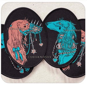 Op de afbeelding: Zwarte ovale patches met gestileerde hondenportretten. Eén hond is roze met een blauwe muilkorf en strik, de andere is turquoise. Beide hebben decoratieve kettingen en de tekst "COEYANDSHY".