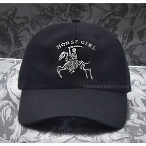 May include: Black baseball cap with a white embroidered design of a skeleton riding a horse. The text "HORSE GIRL" is above the design. The website "COEYANDSHY.COM" is printed on the back of the cap.