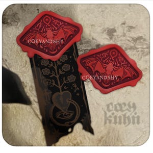 Könnte beinhalten: Zwei rote Wachssiegel mit einem Fledermaus-Design und dem Text "COEYANDSHY". Ein schwarzes Banner mit einem floralen und herzförmigen Design ist ebenfalls sichtbar. Der Hintergrund ist eine strukturierte, cremefarbene Oberfläche.