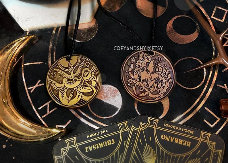 Puede incluir: Dos colgantes de metal dorado en cordones negros. El colgante de la izquierda presenta un dise&ntilde;o detallado de un p&aacute;jaro con una serpiente envuelta a su alrededor. El colgante de la derecha presenta un dise&ntilde;o detallado de una criatura m&iacute;tica con alas y cola. El texto "COEYANDSHY@ETSY" es visible en el fondo.