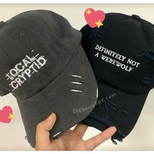 May include: Two black baseball caps with white embroidered text. The first cap says "The Local Cryptid" and the second cap says "Definitely Not A Werewolf".