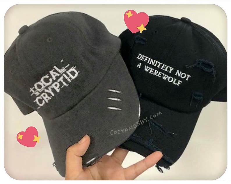 May include: Two black baseball caps with white embroidered text. The first cap says "The Local Cryptid" and the second cap says "Definitely Not A Werewolf".