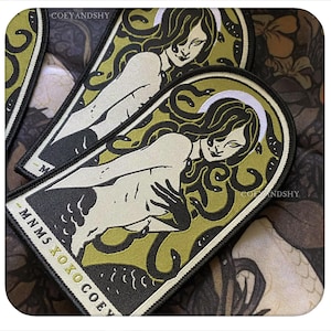 May include: An embroidered patch featuring a stylized figure with snake-like hair, a white halo, and dark accents on an olive green background. The patch has a black border and includes the text MNMS XOXO COEY printed in white and gold lettering.