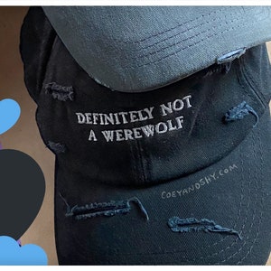 May include: Black distressed baseball cap with the text "DEFINITELY NOT A WEREWOLF" embroidered in white. The cap has a gray underbrim and the website "coeyandshy.com" is printed on the side.