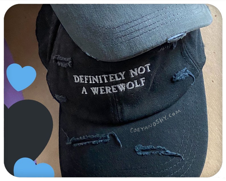 May include: Black distressed baseball cap with the text "DEFINITELY NOT A WEREWOLF" embroidered in white. The cap has a gray underbrim and the website "coeyandshy.com" is printed on the side.