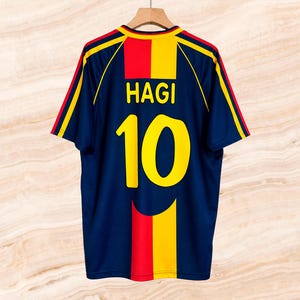 May include: A navy blue short-sleeved sports jersey featuring the name HAGI and the number 10 in yellow on the back. The shirt includes vertical red and yellow stripes down the centre and yellow and red accents on the shoulders and sleeves.
