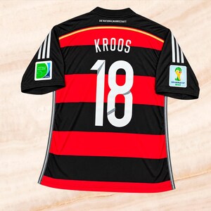 Könnte beinhalten: Ein schwarz-rot gestreiftes Fußballtrikot mit dem Namen KROOS und der Nummer 18 in Weiß auf der Rückseite. Das Trikot hat weiße Streifen an den Ärmeln, FIFA-Weltmeisterschafts-Aufnäher und den Schriftzug DIE NATIONALMANNSCHAFT.