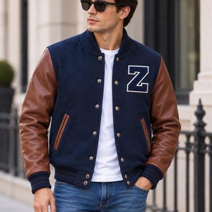 Könnte beinhalten: Eine marineblaue Collegejacke mit braunen Lederärmeln, einem weißen Buchstaben-N-Aufnäher auf der Brust, einem weißen T-Shirt darunter und einer blauen Jeans. Die Jacke verfügt über eine Druckknopfleiste, gerippte Bündchen und zwei Seitentaschen.