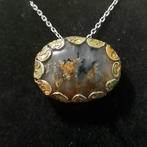 Puede incluir: Un collar con colgante ovalado que presenta una piedra translúcida de tonos marrones y grises moteados con inclusiones, engastada en un bisel de metal dorado texturizado y festoneado, suspendido de una fina cadena plateada.