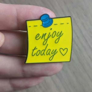 May include: A square yellow enamel pin shaped like a sticky note with a black border. The pin features the text "enjoy today" in black cursive script, a small black heart symbol, and a blue thumbtack graphic at the top center.