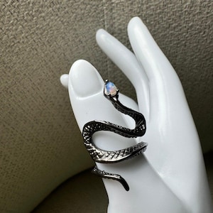 May include: A dark metallic snake-shaped ring featuring a textured, scale-like pattern along its body. The head of the snake is adorned with a small, oval-shaped iridescent stone that displays subtle blue and white light reflections.