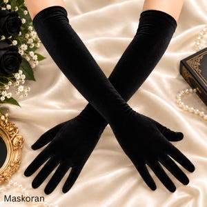 Peut inclure: Une paire de longs gants noirs en velours de style opéra qui dépassent le coude. Les gants présentent une texture lisse et mate ainsi qu'une coupe ajustée couvrant les doigts, les paumes et les avant-bras, avec le texte Maskoran en bas.
