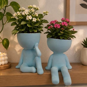 May include: Two light blue ceramic planters shaped like seated figures. The left planter holds a plant with small white flowers, while the right planter holds a plant with small pink flowers. Both figures have smooth, rounded heads and limbs.