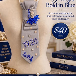 May include: A gray necktie featuring a pearl necklace collar, adorned with blue crystal accents, the text 1920, and the script text Zeta Phi. The tie is decorated with scattered pearl and rhinestone embellishments, displayed on a mannequin torso.