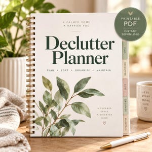 Può includere: Un'agenda Declutter Planner a spirale con copertina color crema decorata con foglie di eucalipto verdi. Il testo riporta A Calmer Home A Happier You, Declutter Planner, Plan Sort Organize Maintain e A Cleaner Space A Brighter Mind con un cuore.