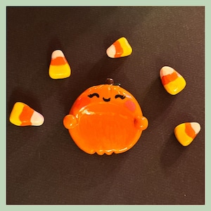 May include: A glossy orange pumpkin-shaped dish with a painted smiling face, closed eyes, and pink cheeks, surrounded by five individual candy corn pieces featuring white, orange, and yellow horizontal stripes arranged in a circular pattern.