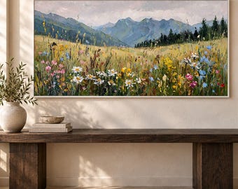 May include: A rectangular landscape painting in a white frame depicting a mountain range behind a field of wildflowers. The scene features yellow, white, and pink flowers in the foreground with green hills and dark trees under a cloudy sky.